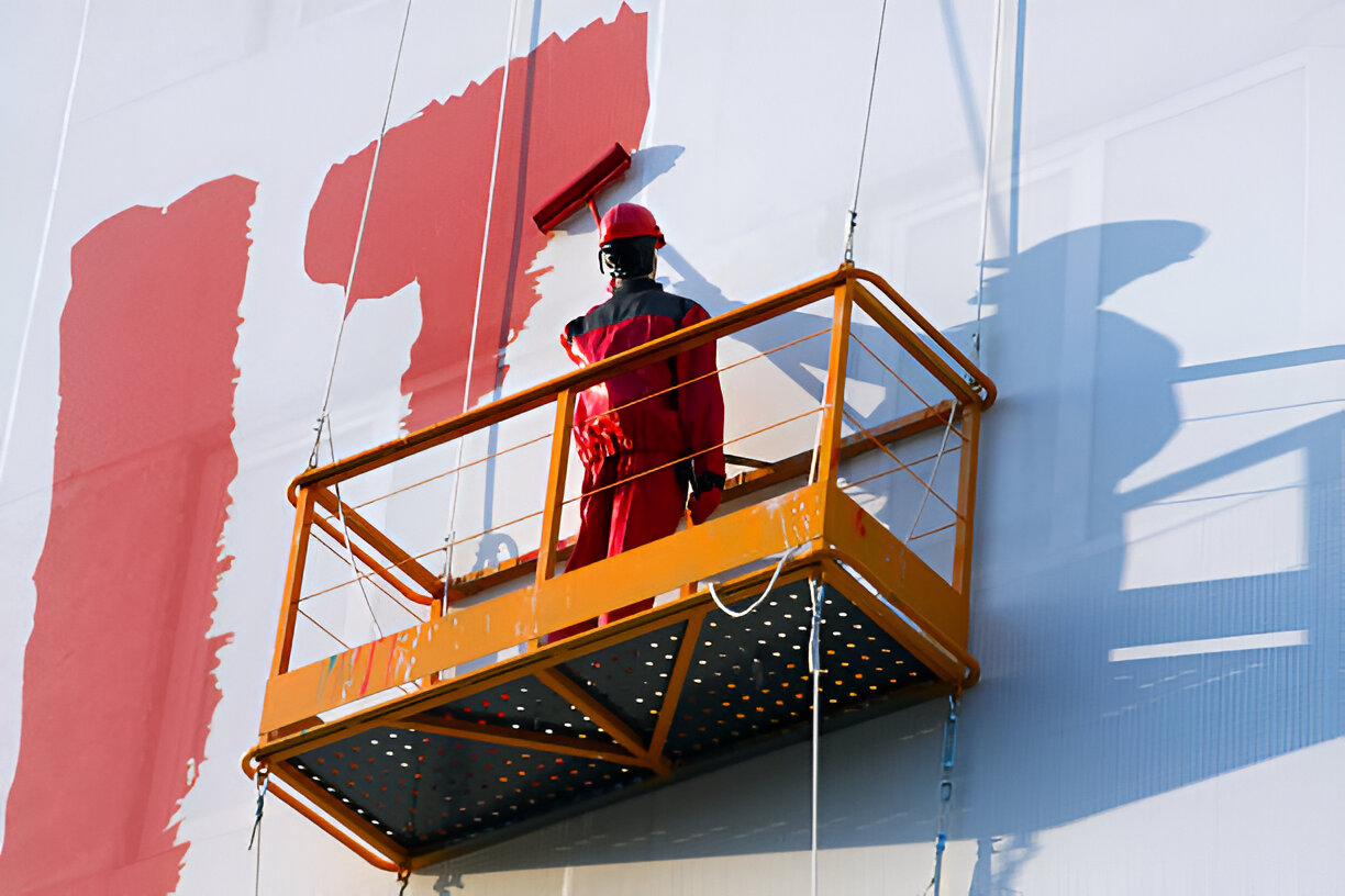 Worker in red protective gear painting a large industrial structure with a roller on an elevated platform, under bright daylight
